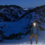 hiker with a headlamp standing on a snow covered mountain slope at dusk using trekking poles and carrying a backpack