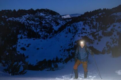 Hiker with a headlamp standing on a snow-covered mountain slope at dusk, using trekking poles and carrying a backpack.
