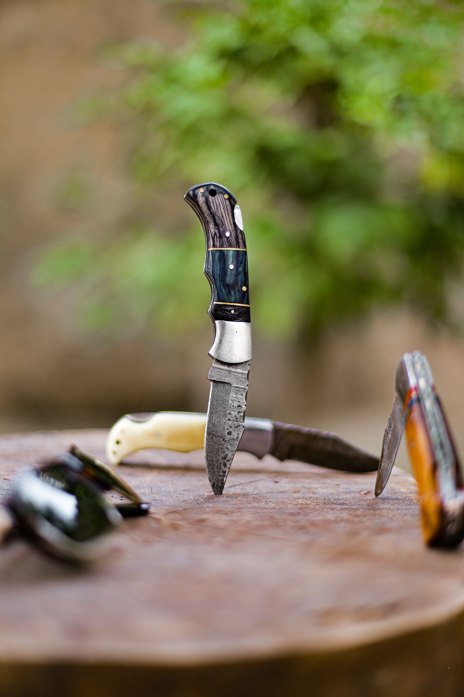 several knives with ornate handles stand upright in a weathered wooden surface outdoors with a blurred green background